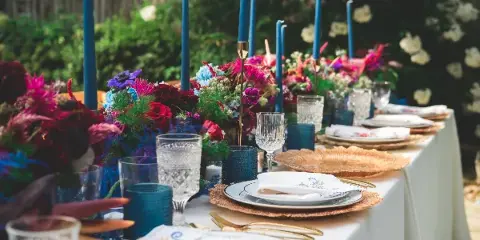 A close-up view of a wedding table setting featuring white plates, gold flatware, crystal glassware, blue candle holders, and a vibrant floral centerpiece.