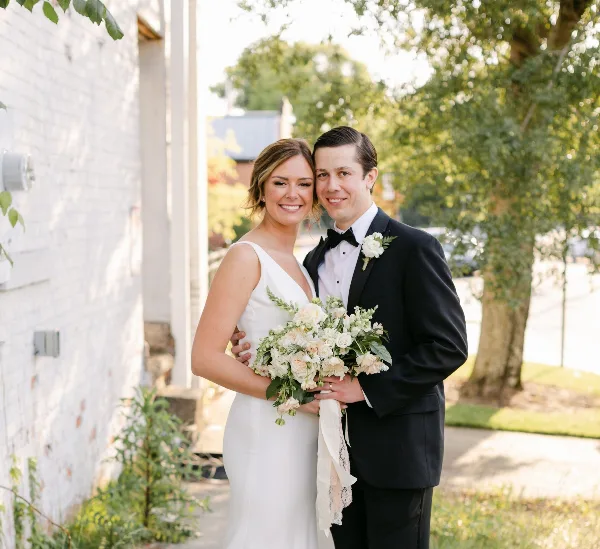 A bride and groom posing together outdoors, with the bride holding a bouquet of white and cream flowers.