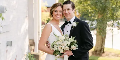 A bride and groom posing together outdoors, with the bride holding a bouquet of white and cream flowers.