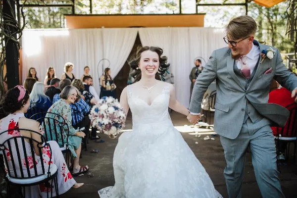 A bride and groom walking down the aisle during a wedding ceremony at a semi-outdoor venue with white drapery and guests seated on chairs.