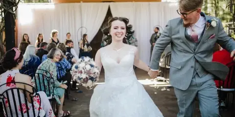 A bride and groom walking down the aisle during a wedding ceremony at a semi-outdoor venue with white drapery and guests seated on chairs.