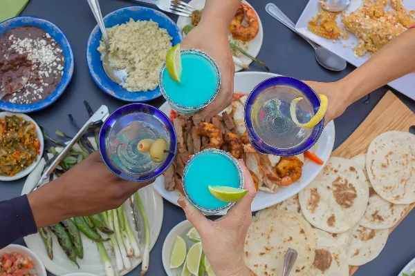 A top-down view of a table filled with Southwestern dishes including tortillas, rice, and grilled meats, with four hands toasting with blue margaritas.