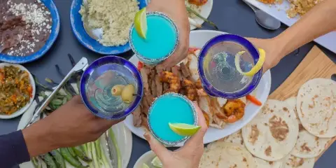A top-down view of a table filled with Southwestern dishes including tortillas, rice, and grilled meats, with four hands toasting with blue margaritas.