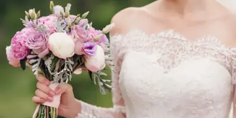 A bride in a white lace wedding dress holding a bouquet of pink and white flowers.