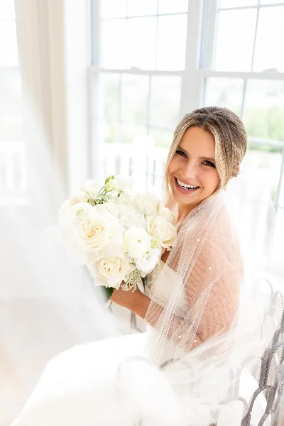 A smiling bride with styled hair and soft makeup holding a white rose bouquet in a bright room.