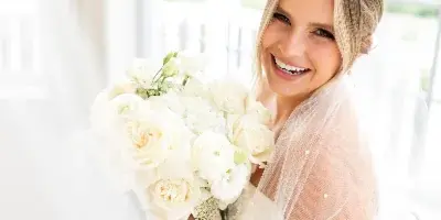 A smiling bride with styled hair and soft makeup holding a white rose bouquet in a bright room.