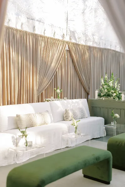 A professionally designed wedding lounge area featuring a white sofa, green velvet bench, and floral arrangements against a backdrop of beige drapery.