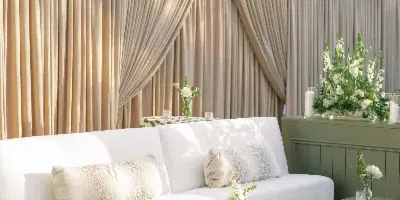A professionally designed wedding lounge area featuring a white sofa, green velvet bench, and floral arrangements against a backdrop of beige drapery.