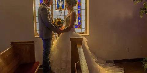A bride and groom standing in a church aisle in front of a stained glass window.