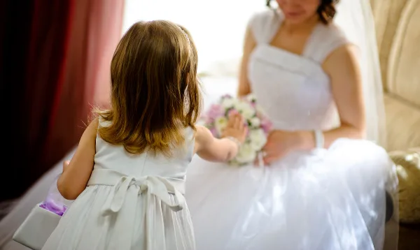 A young girl in a white dress reaches out to a bride holding a floral bouquet.