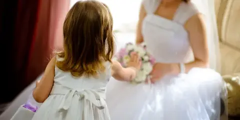A young girl in a white dress reaches out to a bride holding a floral bouquet.
