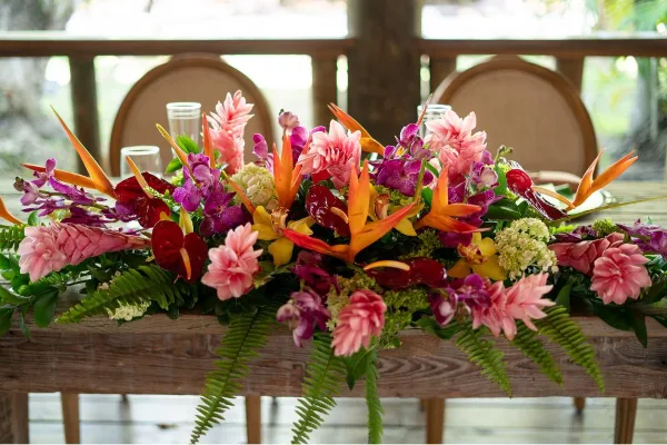 A vibrant floral centerpiece featuring pink ginger, orange bird of paradise, and purple orchids arranged on a wooden table.