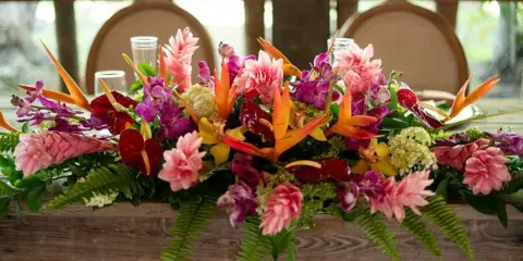 A vibrant floral centerpiece featuring pink ginger, orange bird of paradise, and purple orchids arranged on a wooden table.