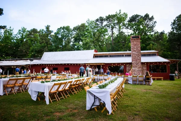 Outdoor wedding reception at a rustic red barn venue with long tables, wooden chairs, and string lights.