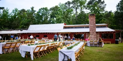 Outdoor wedding reception at a rustic red barn venue with long tables, wooden chairs, and string lights.