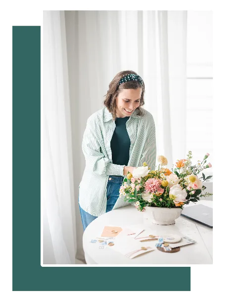 A woman in a light green shirt and headband smiles while arranging a floral centerpiece and reviewing wedding stationery on a white table.