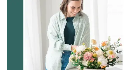 A woman in a light green shirt and headband smiles while arranging a floral centerpiece and reviewing wedding stationery on a white table.