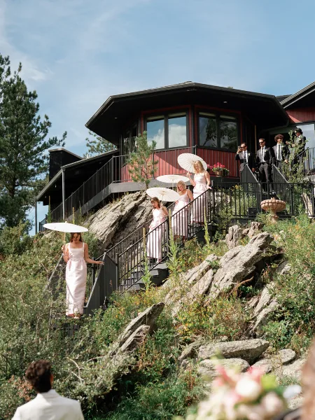 Bridesmaids in light dresses descend a rocky outdoor staircase with parasols near a red and black lodge-style wedding venue.