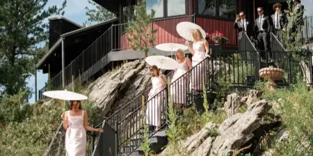 Bridesmaids in light dresses descend a rocky outdoor staircase with parasols near a red and black lodge-style wedding venue.