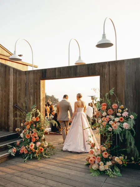 A bride and groom walking through a wooden doorway decorated with large floral arrangements at a wedding venue.