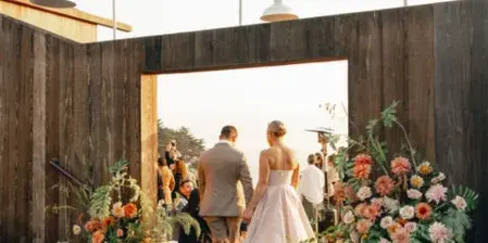 A bride and groom walking through a wooden doorway decorated with large floral arrangements at a wedding venue.