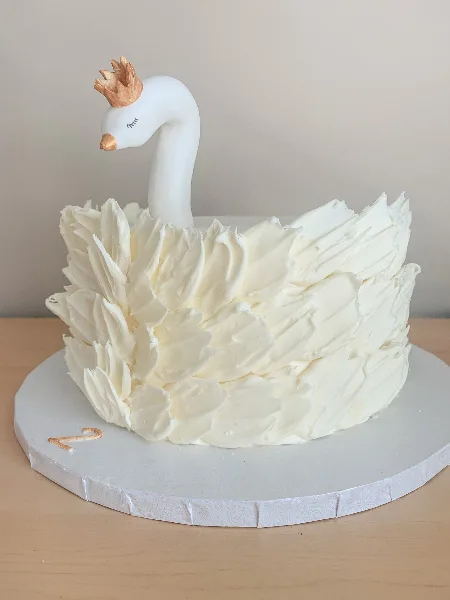 A white custom cake decorated with textured buttercream petals and a sculpted white swan topper with a gold crown.