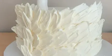 A white custom cake decorated with textured buttercream petals and a sculpted white swan topper with a gold crown.