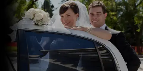 A bride and groom smiling from a white limousine decorated with a floral arrangement on the roof.