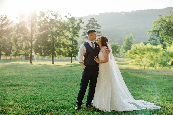 A bride and groom embracing in a grassy field with trees and hills in the background.
