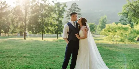 A bride and groom embracing in a grassy field with trees and hills in the background.