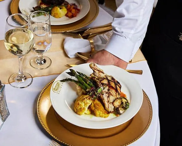 A server in a white chef's coat places a plated dish of grilled chicken, asparagus, and cauliflower on a gold-rimmed plate at a formally set event table with wine glasses.