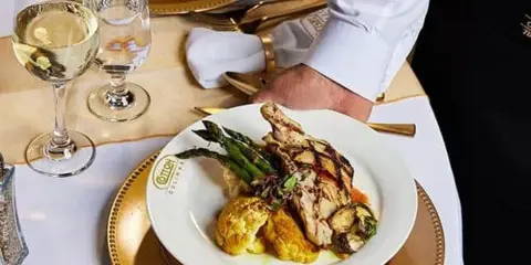 A server in a white chef's coat places a plated dish of grilled chicken, asparagus, and cauliflower on a gold-rimmed plate at a formally set event table with wine glasses.