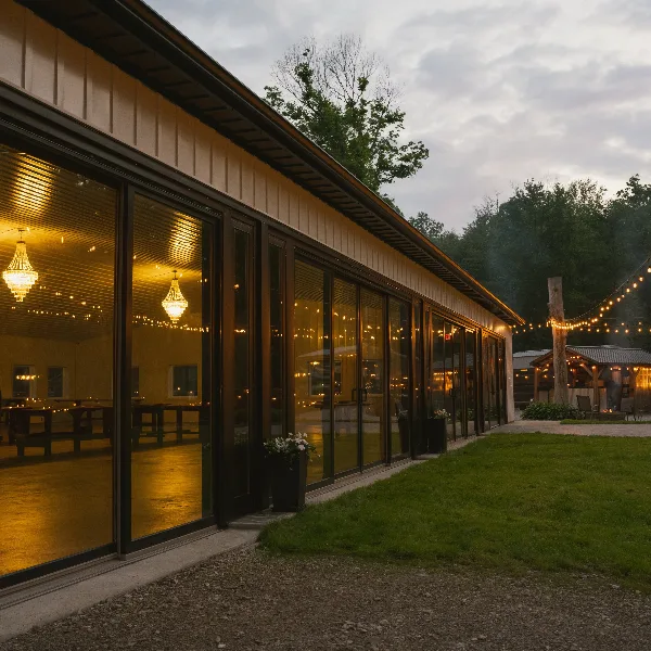 Exterior view of a modern glass-walled wedding reception hall with warm interior lighting and string lights at dusk.