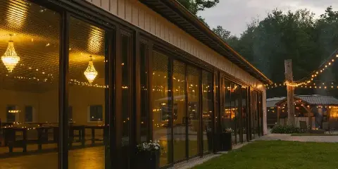 Exterior view of a modern glass-walled wedding reception hall with warm interior lighting and string lights at dusk.