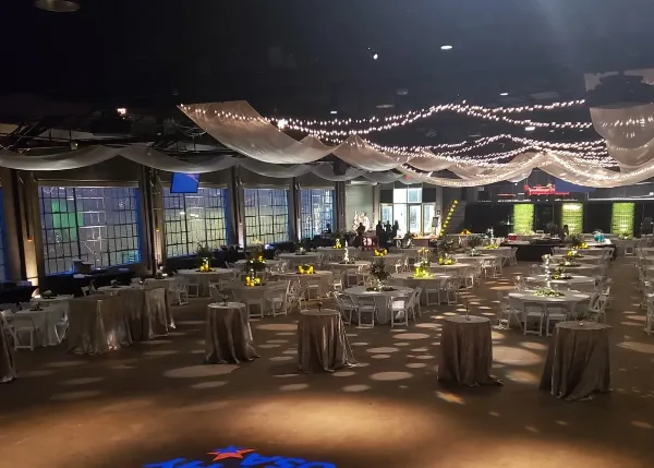 An indoor event space set up with round tables, white chairs, and draped ceiling fabric with string lights.