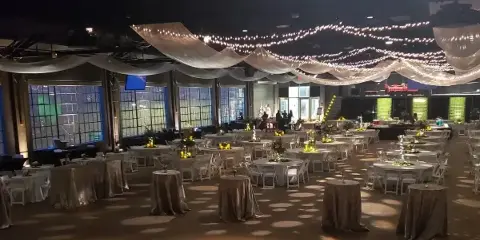 An indoor event space set up with round tables, white chairs, and draped ceiling fabric with string lights.