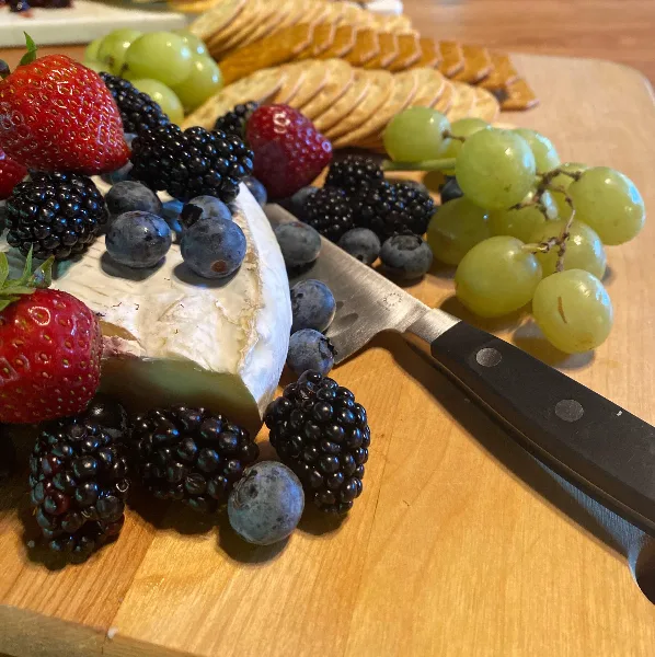 A catering platter featuring a wedge of soft cheese, fresh strawberries, blackberries, blueberries, green grapes, and crackers arranged on a wooden board.