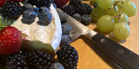 A catering platter featuring a wedge of soft cheese, fresh strawberries, blackberries, blueberries, green grapes, and crackers arranged on a wooden board.