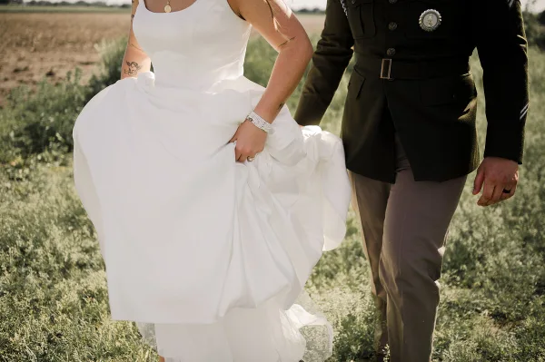 A bride in a white dress and a groom in a military uniform walking together in a grassy field.