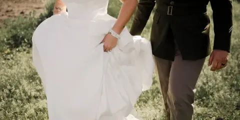 A bride in a white dress and a groom in a military uniform walking together in a grassy field.