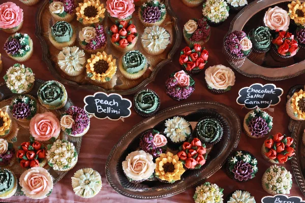 A collection of colorful, flower-decorated cupcakes arranged on silver serving platters with handwritten flavor labels.