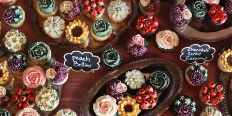 A collection of colorful, flower-decorated cupcakes arranged on silver serving platters with handwritten flavor labels.