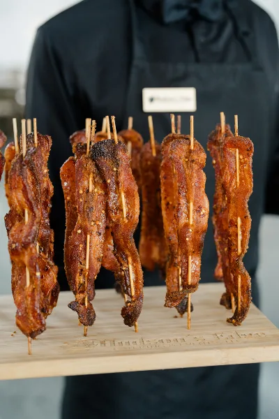 A catering staff member in a black apron holding a wooden board with skewers of glazed bacon, branded with the text 'Hudson's Edge Catering & Events'.