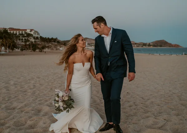 A bride in a white gown and a groom in a navy suit walking hand-in-hand on a sandy beach at dusk.