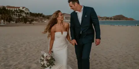 A bride in a white gown and a groom in a navy suit walking hand-in-hand on a sandy beach at dusk.