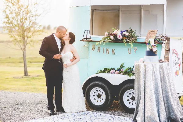 A bride and groom kiss next to a vintage teal rental truck decorated with flowers and a 'DRINK UP' sign, with a decorated table nearby.