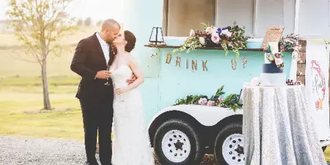A bride and groom kiss next to a vintage teal rental truck decorated with flowers and a 'DRINK UP' sign, with a decorated table nearby.