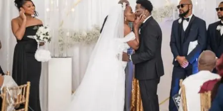 Bride and groom sharing a kiss during a wedding ceremony at an indoor venue with a floral arch and seated guests.