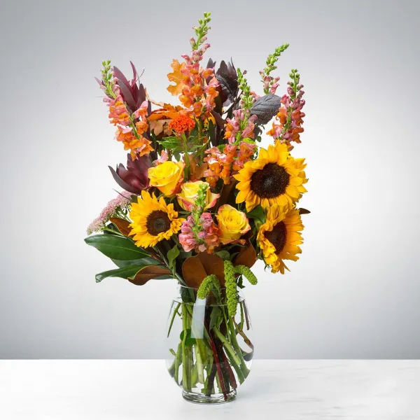 A tall floral arrangement featuring sunflowers, yellow roses, and snapdragons in a clear glass vase.