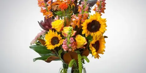 A tall floral arrangement featuring sunflowers, yellow roses, and snapdragons in a clear glass vase.